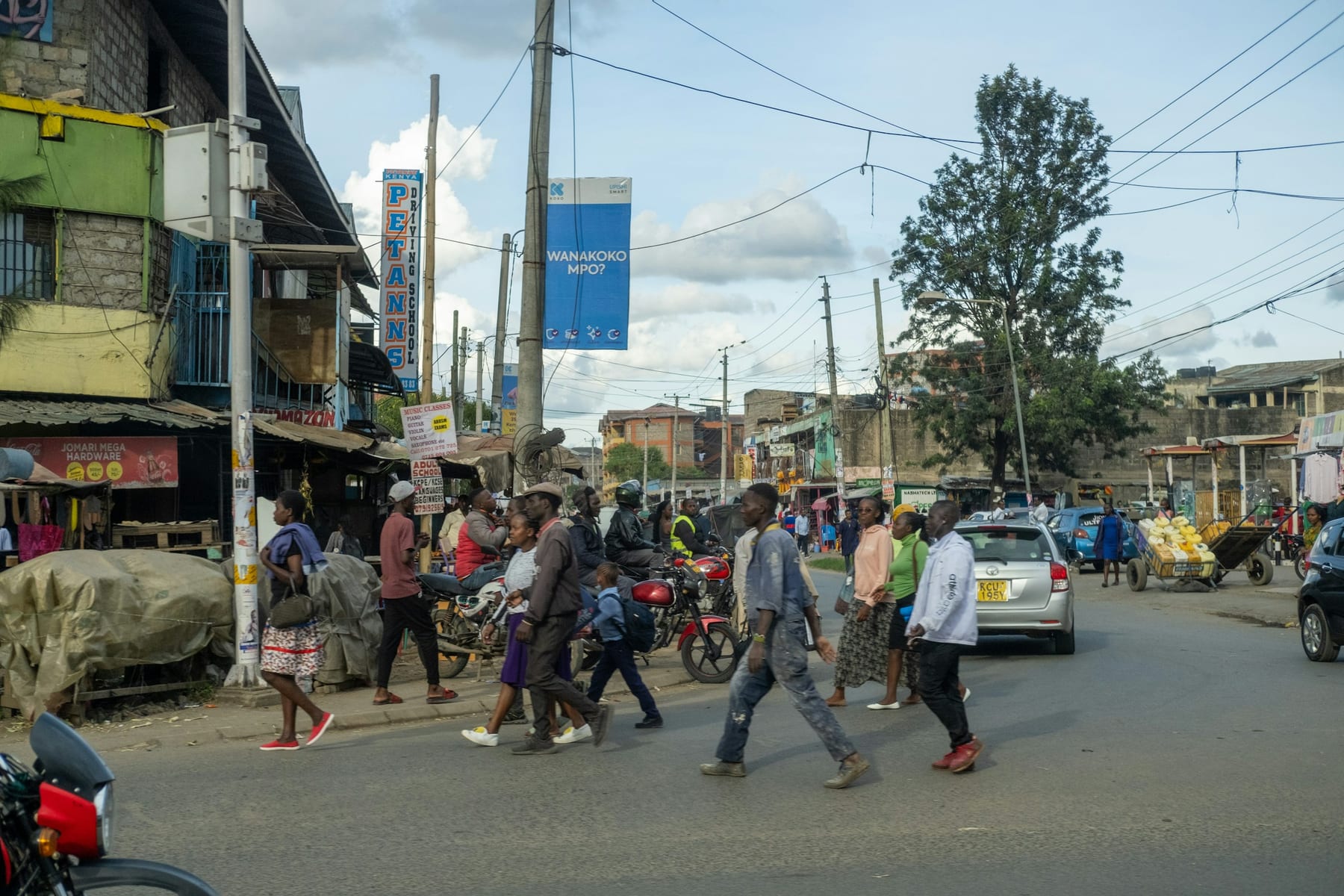 A busy Nairobi street corner, people crossing between the shops and the stage.