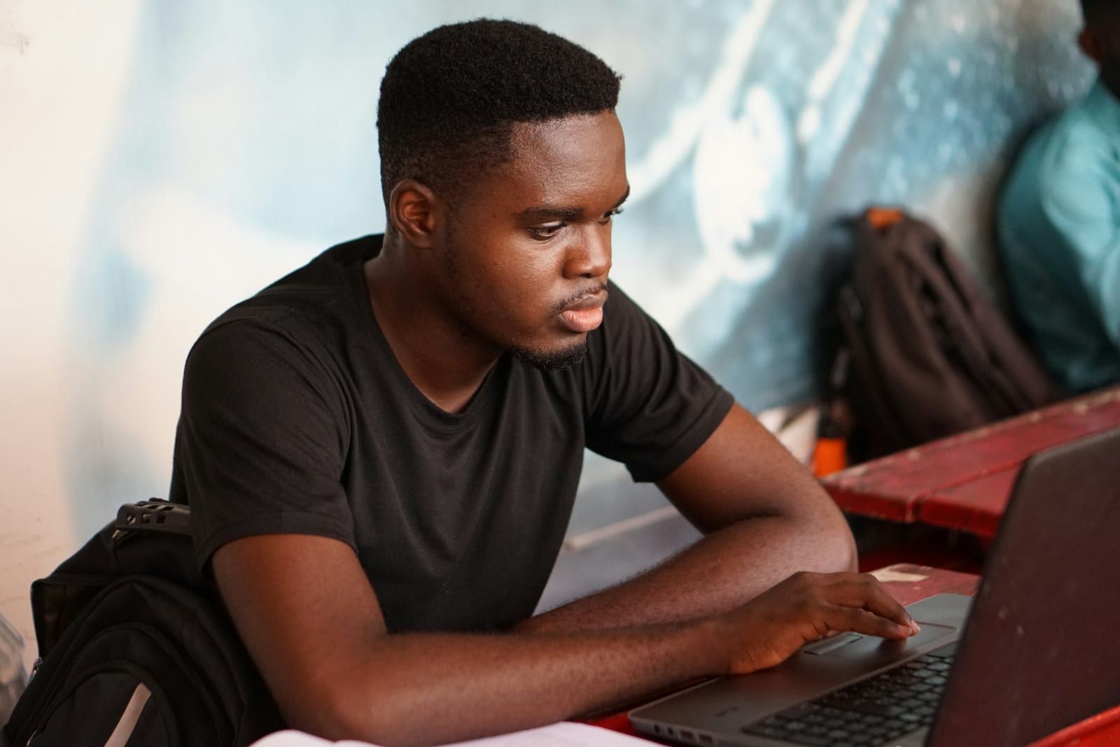 A student working at a laptop, a backpack beside him.